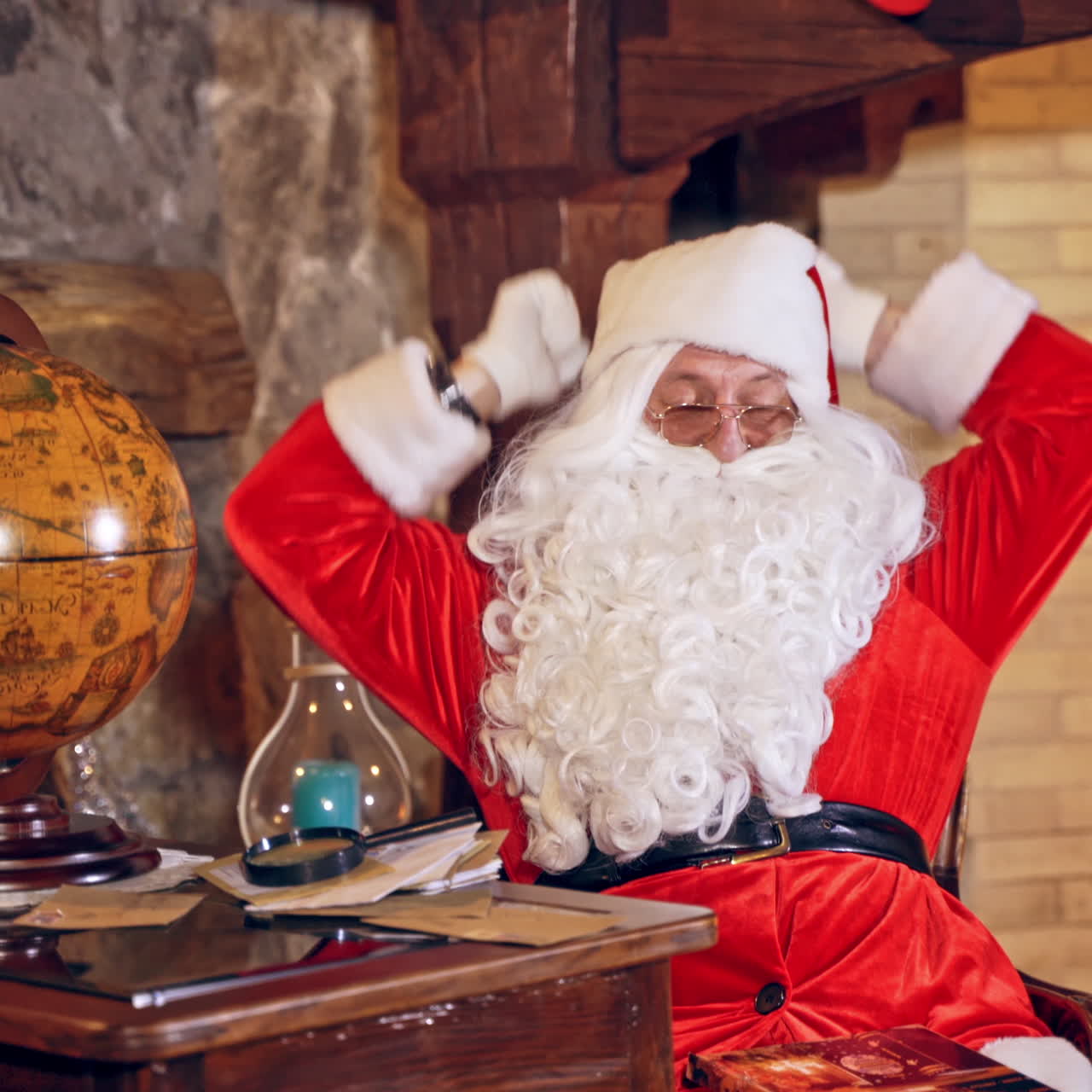 Santa Claus is sitting in specially decorated room at the table, holding an envelope in hands. Feast fireplace in the background. Christmas spirit concept close up video. Christmas eve. New Year is coming. Santa Claus is looking at the envelope with a magnifying glass.