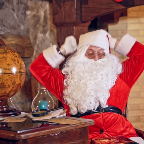santa claus is sitting in specially decorated room at the table, holding an envelope in hands. feast fireplace in the background. christmas spirit concept close up video. christmas eve. new year is coming. santa claus is looking at the envelope with a magnifying glass.