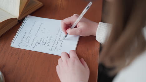 partial view of lady writing on spiral notebook using transparent pen beside open novel and coffee cup on wooden desk in cozy indoor space with natural light