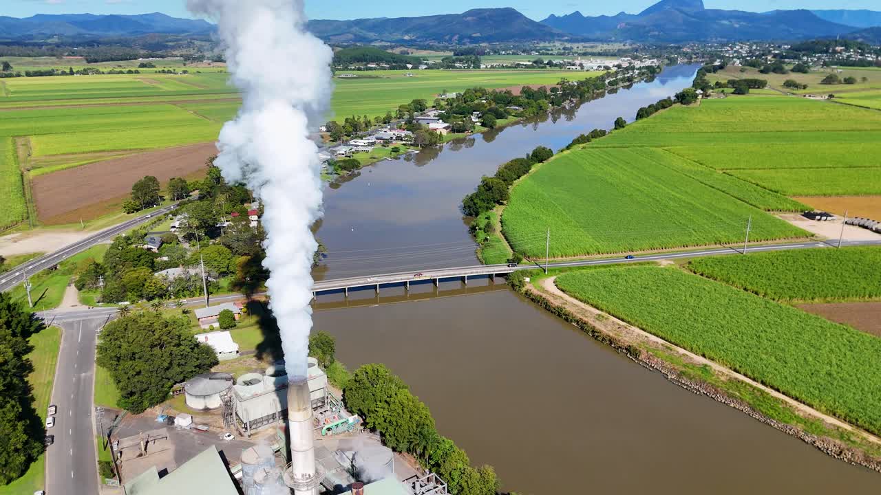 Drone footage captures steam rising from a factory amidst lush green fields and a winding river under clear skies