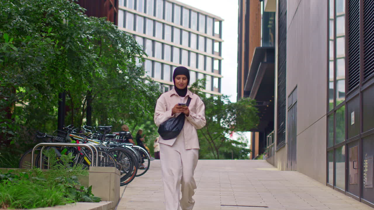 mujer de negocios musulmana con hijab yendo a trabajar de pie fuera de una oficina moderna mirando su teléfono móvil 1