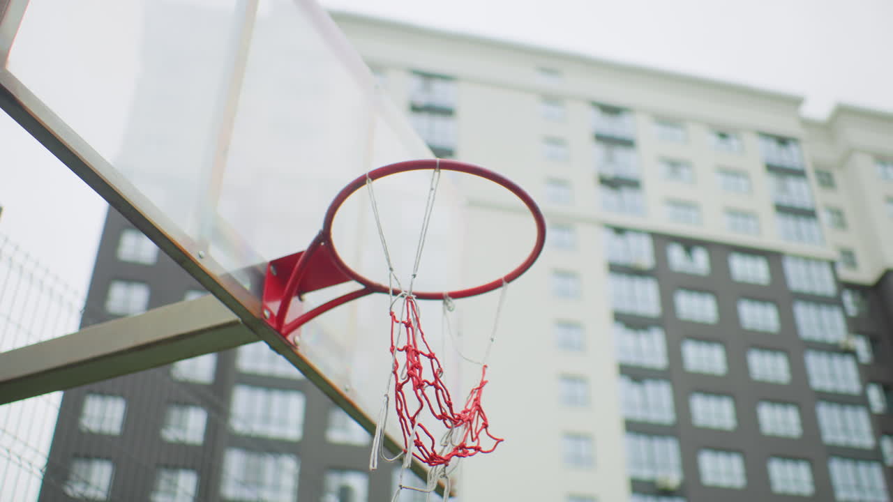 Canestro da basket urbano sopra un canestro di metallo, primo piano del cerchio rosso e della rete in nylon con grattacieli sullo sfondo, ripresa al rallentatore della palla che cade a segno, energia della partita improvvisata, esultanza del giovane giocatore