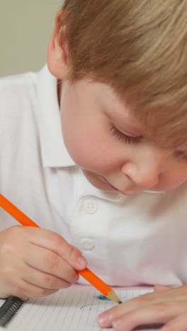 blond toddler examines pencil sitting at table in cozy apartment. cute boy draws picture in notebook carefully. obedient child studies closeup