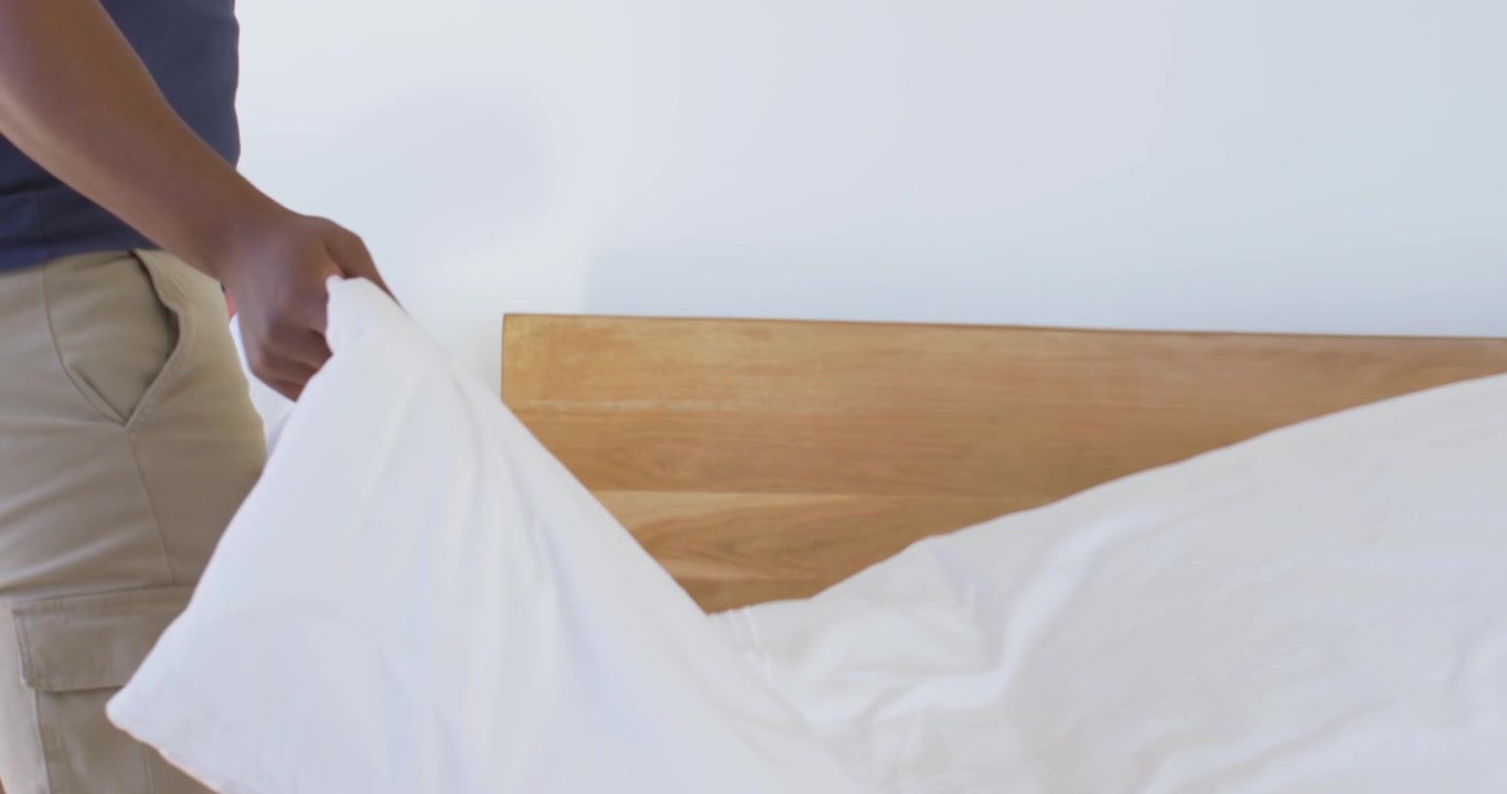 African American man holding and shaking white duvet at bedside, making bed, tidying, copy space
