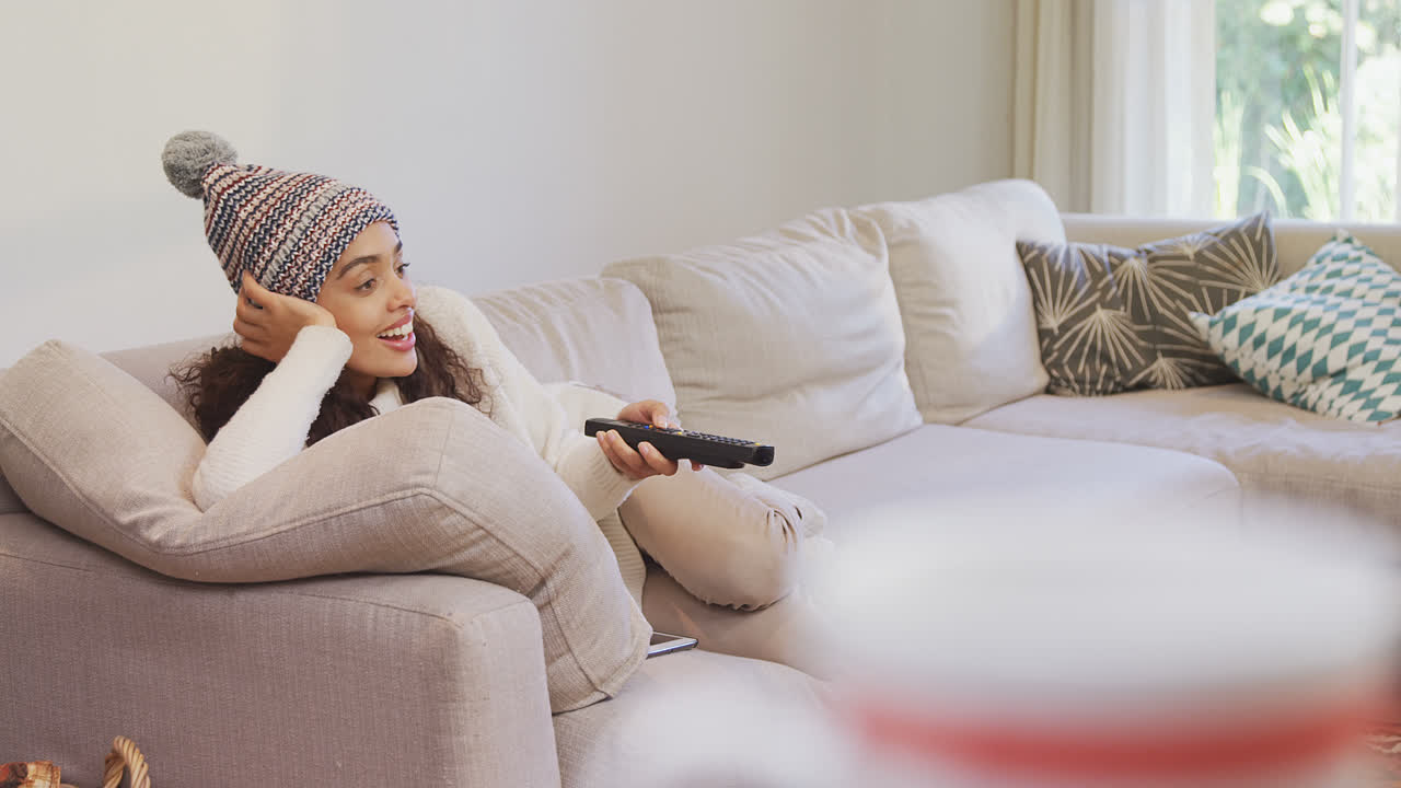 따뜻한 옷을 입은 행복한 여성이 소파에 누워서 텔레비전 4k 4k를 보고 있습니다.