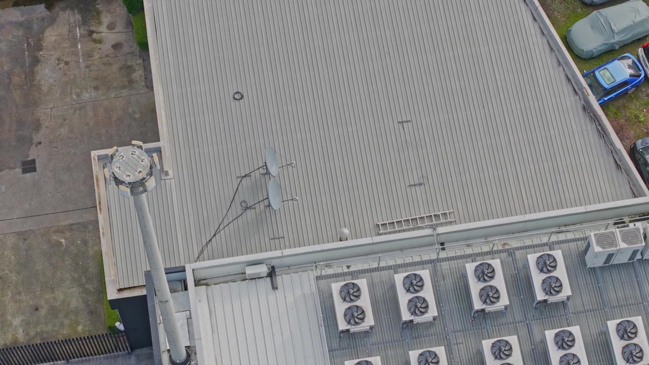 Drone shot of a data centre rooftop reveals large cooling units, satellite antennas, and utility poles. Bright daylight provides clear detail on metallic surfaces, advanced infrastructure.