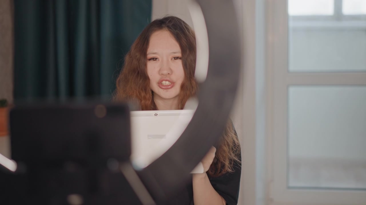 Una giovane donna in uno studio casalingo tiene e regola il tablet mentre si gira verso la fotocamera, a metà azione con la testa girata e un'espressione facciale concentrata che indica una dimostrazione o spiegazione attiva.