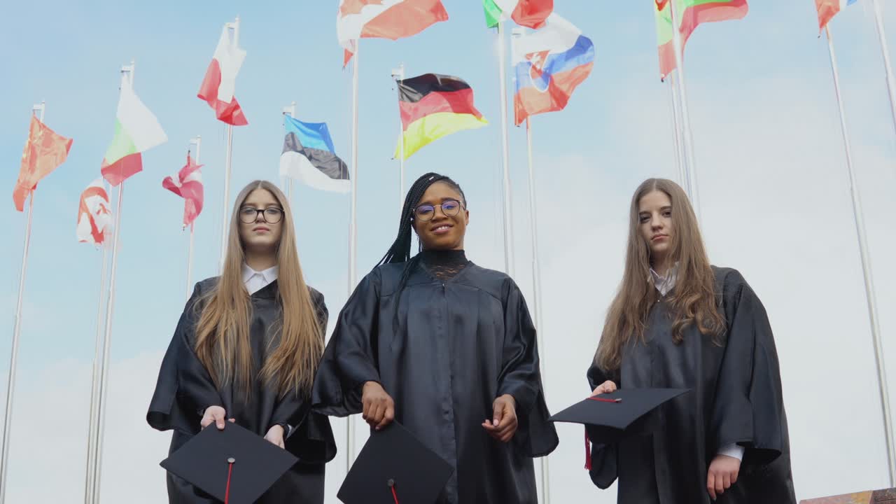 Three young women of different nationalities joyfully throw hats up against many flags of different countries fluttering in the wind on the background. Outdoor view