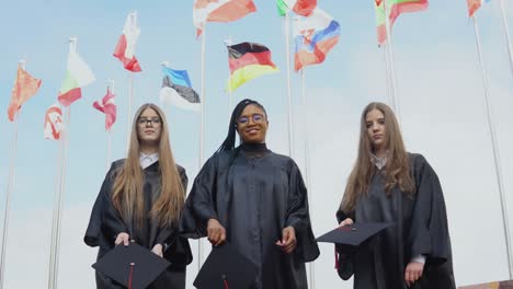 tres mujeres jóvenes de diferentes nacionalidades alegremente lanzan sombreros contra muchas banderas de diferentes países revoloteando en el viento en el fondo. vista al aire libre