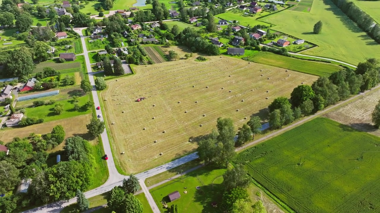 Drone shot over village fields with tractor rolling hay bales in warm sunlight