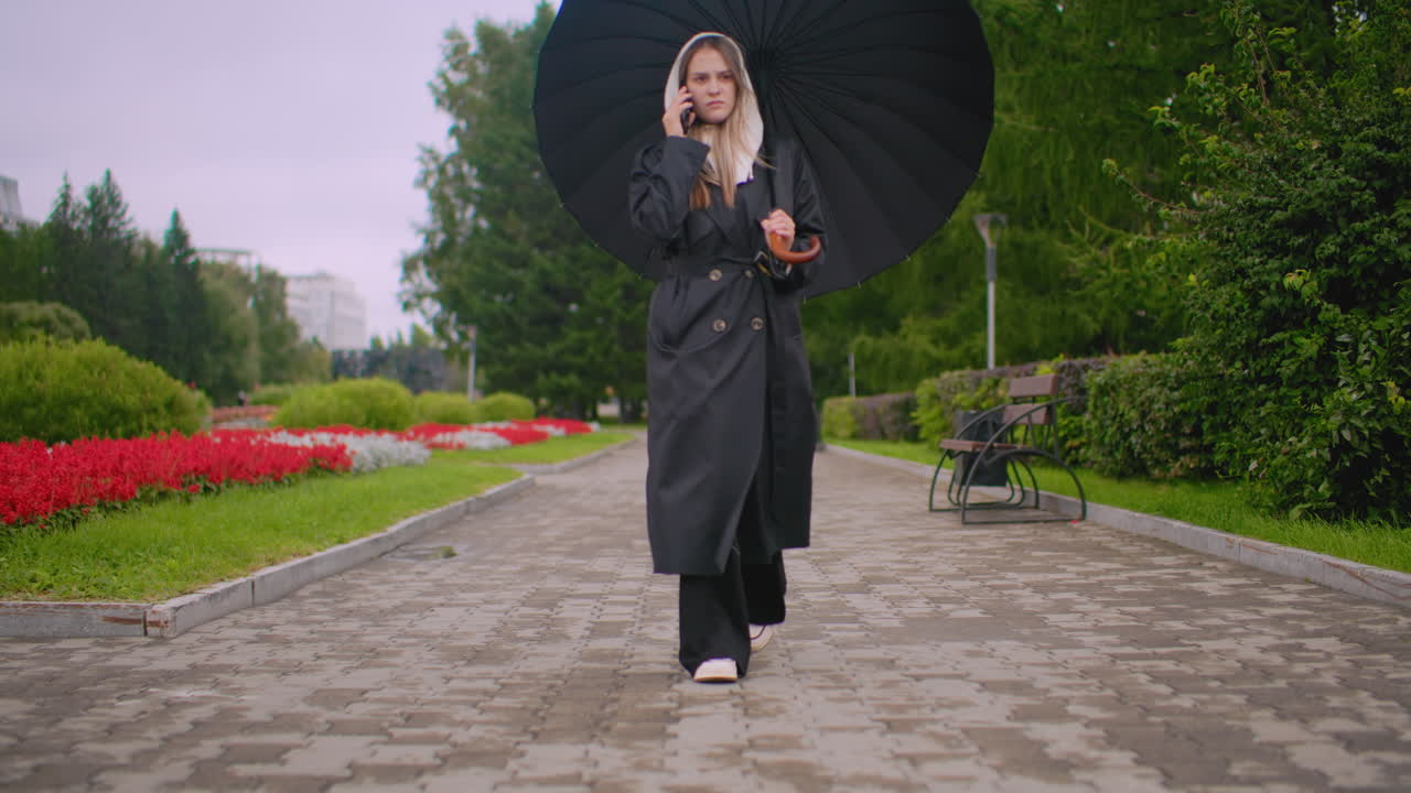 Ragazza con ombrello e cappotto nero cammina su un sentiero di pietra bagnata in un parco mentre parla al telefono durante una giornata piovosa, circondata da fiori rossi, panchine e alberi verdeggianti che creano un'atmosfera urbana serena