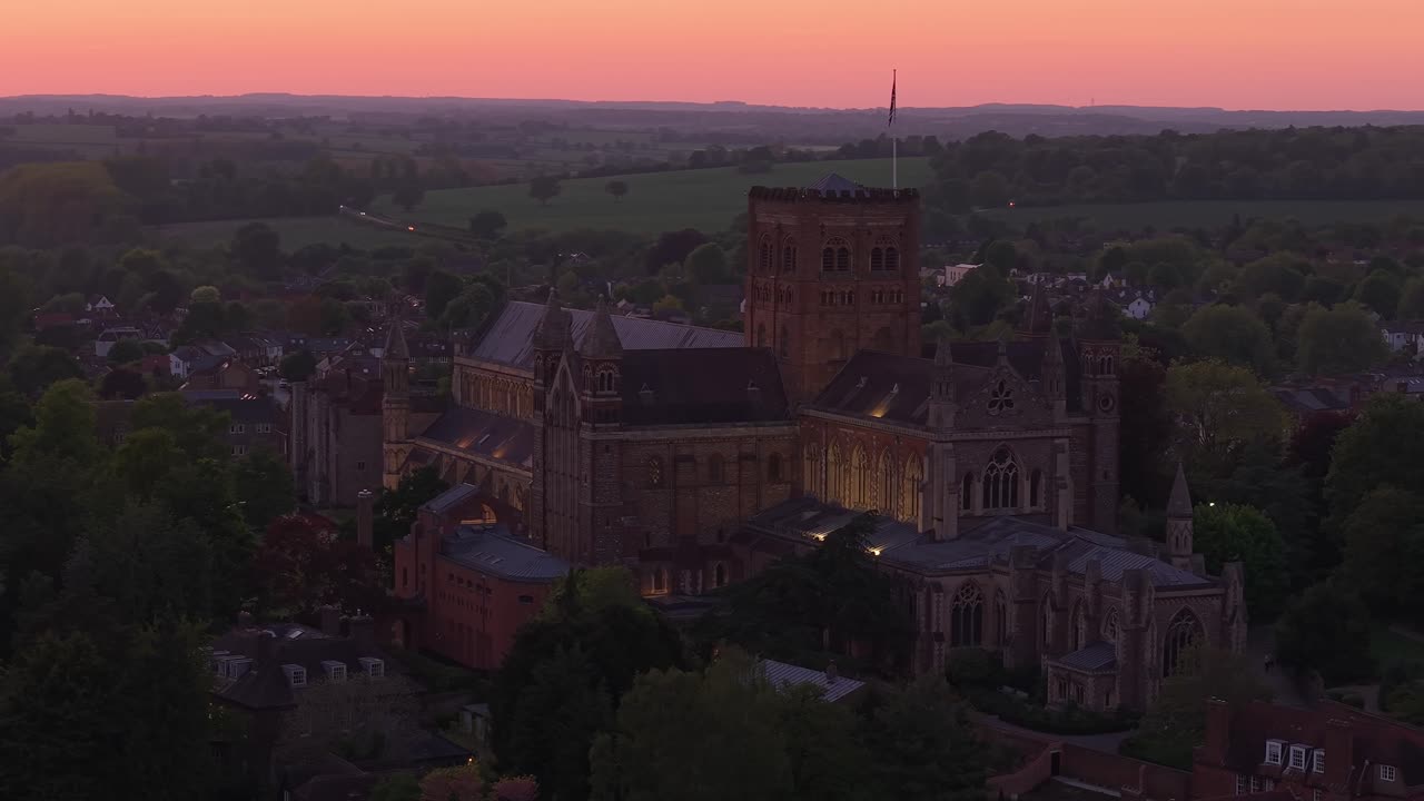 Aerial telephoto drone captures the majestic illuminated St Albans Cathedral at night, highlighting its architectural details and glowing presence against the evening cityscape
