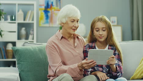 foto de retrato de la abuela de cabello gris y la bonita nieta adolescente sentada en el sofá en la acogedora habitación y viendo algo en la pantalla de la tableta