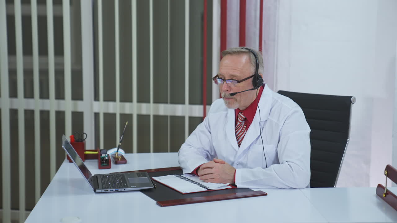Remote medical consultation. Professional male doctor in white medical coat and headset making conference call on laptop. Physician explains treatment by webcam.