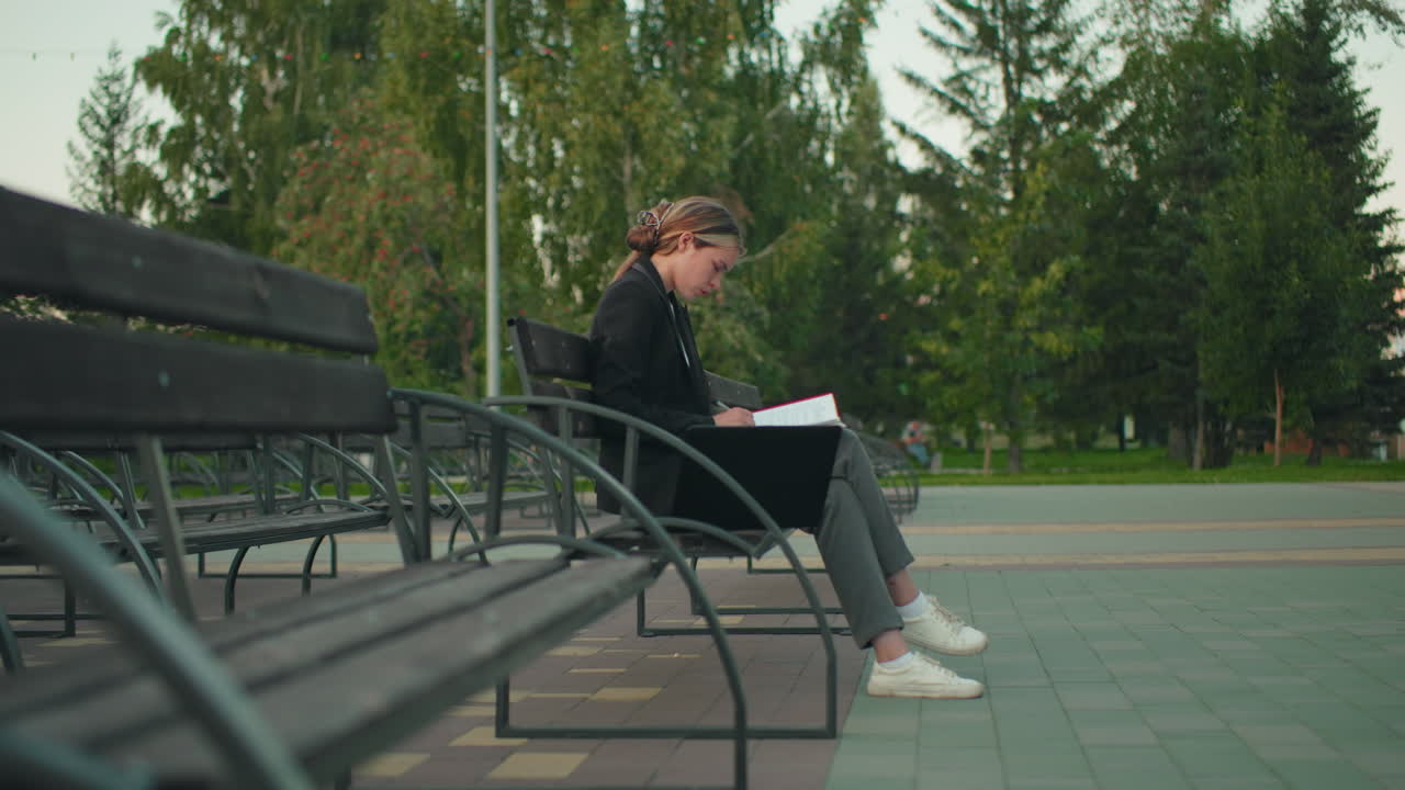 Student in professional attire seated alone on outdoor bench flipping through red folder with pen, laptop beside her, in quiet park with trees and distant people in blur view