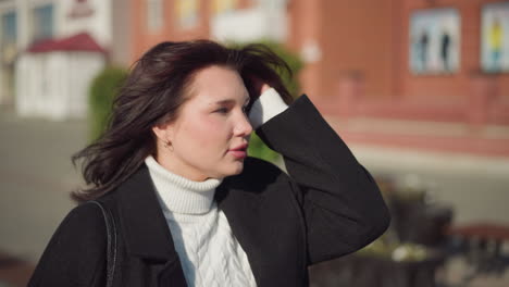 young lady with brown hair in black coat adjusting her hair while looking to her left, dressed in a white cable knit sweater, standing on a sunny urban street