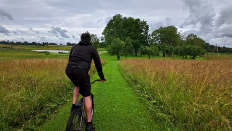 young man riding a bike in a beautiful green field with a view of a cabin on a lake