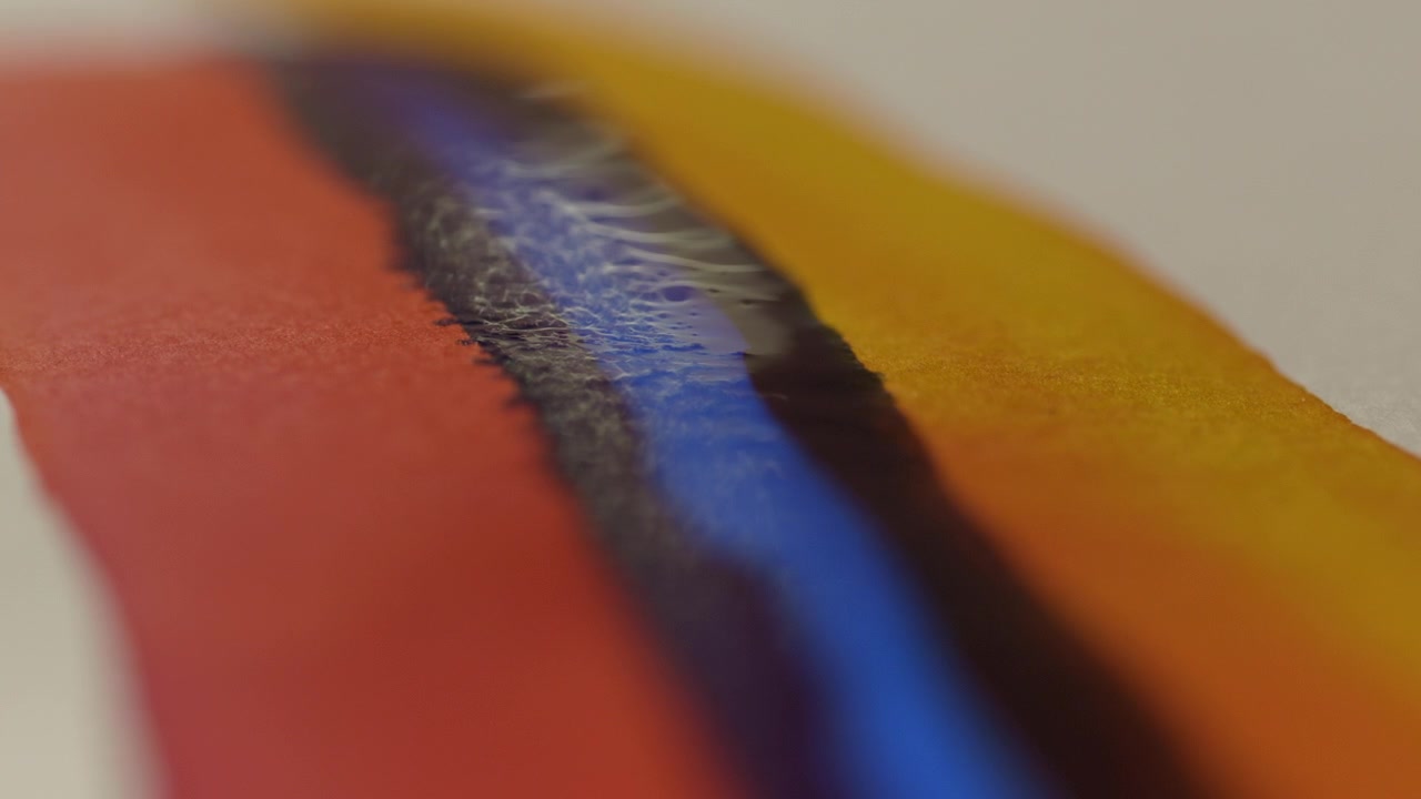 Extreme macro close-up of an artist applying a blue brushstroke over red and yellow paint on paper. Shallow depth of field highlights texture, color layering and creative process.