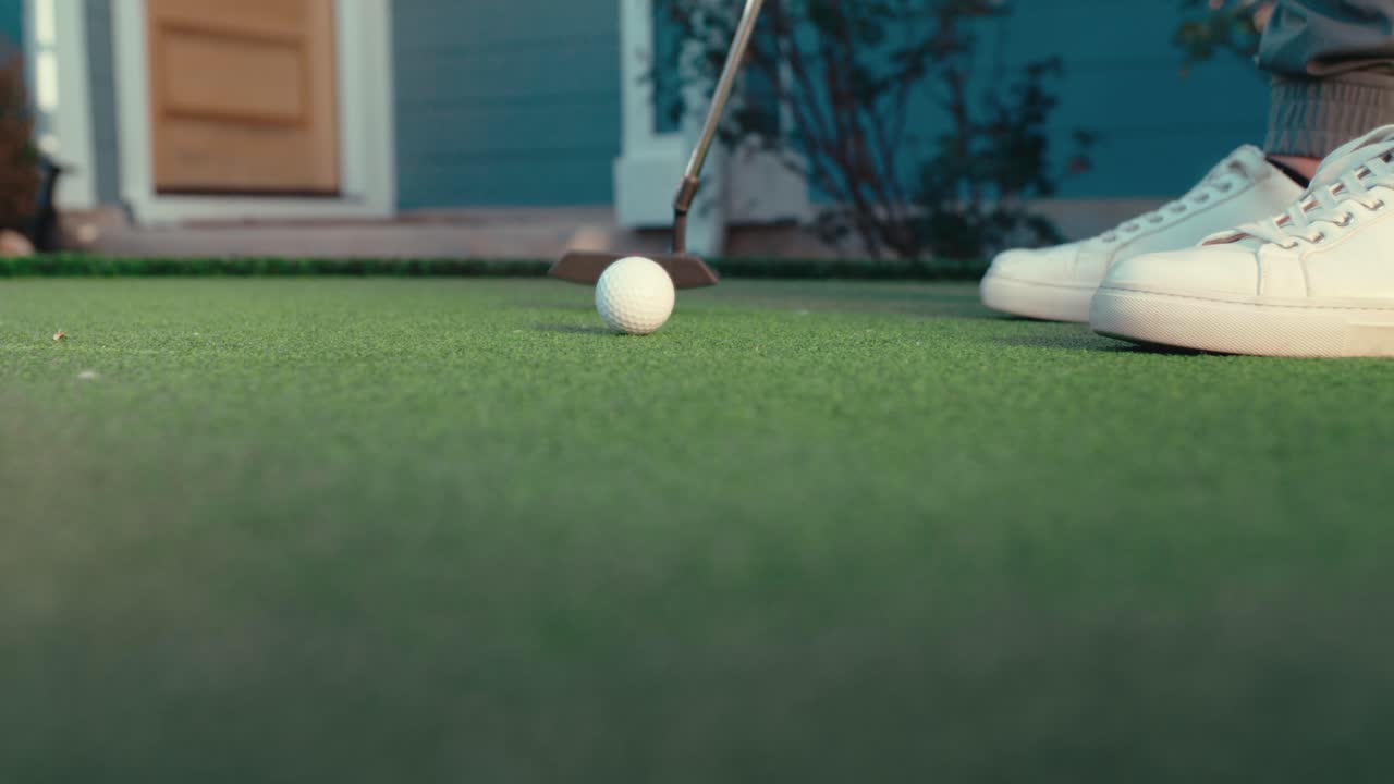 Zpomalený záběr putteru odpalujícího míček směrem ke kameře na venkovním domácím greenu, s vchodem do obytného domu v pozadí. Ideální pro golfová tréninková videa, lifestylový obsah a reklamy na produkty.