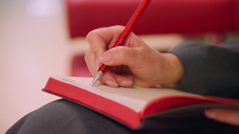 close-up of person holding red pen, writing in notebook with soft-focus background. creative and professional business planning, journaling, or note-taking action in modern workspace