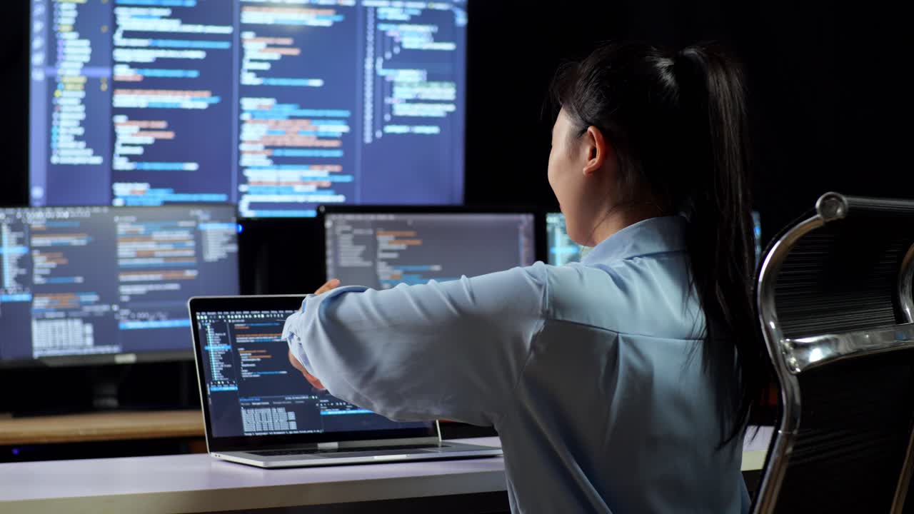 vista trasera de una programadora asiática estirándose mientras escribe código en una computadora portátil usando múltiples monitores que muestran la base de datos en los escritorios de ventana de terminal en la oficina