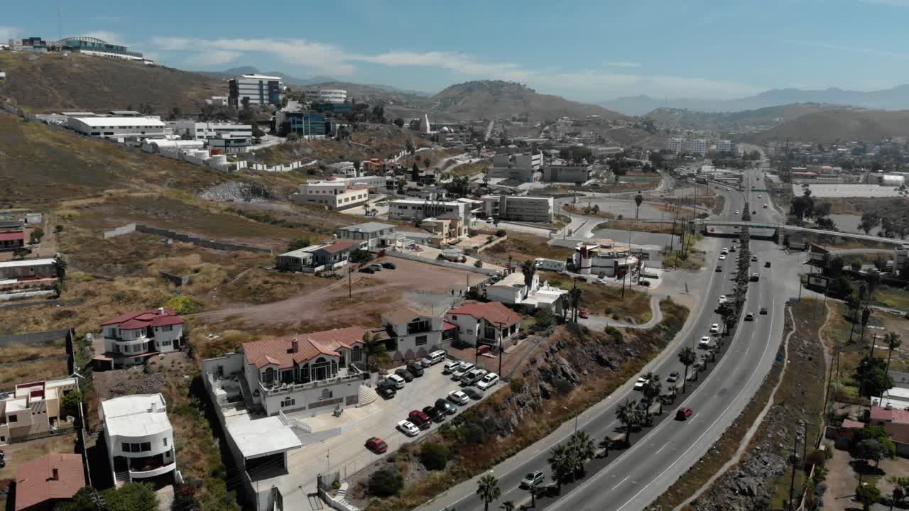 Ripresa aerea con drone di un'area residenziale collinare con case, strade e auto parcheggiate, terreno arido ed edifici sparsi che mostrano lo sviluppo suburbano e una vista panoramica dall'alto.