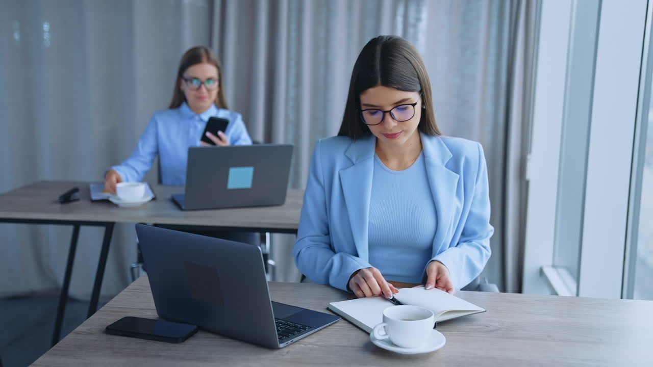 Young ladies working in the office on laptops. Brunette woman looks at the screen and Blonde one drinking coffee and looking at phone. Blurred backdrop.