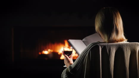 woman with a book and a glass of wine is relaxing at home by the fireplace