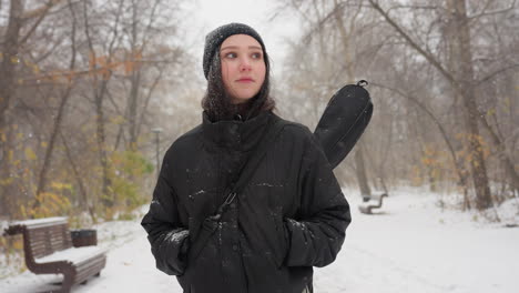 girl in black hoodie carrying guitar in pack looks around thoughtfully while snow falls gently, with a snow-covered bench in the background enhancing the wintry scene