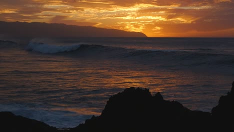 una gran ola llega cuando el sol acaba de ponerse sobre la costa norte de hawaii con rocas volcánicas en la playa recortadas contra la luz naranja