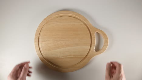 top view: human hands puts a round cutting board with handly on a white table