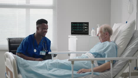 a black cardiologist doctor is talking to a 60-70-year-old patient lying on a bed in a hospital. a neurologist is talking to a patient. the patient is connected to an oxygen mask and an ecg device