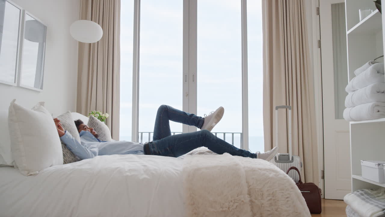 행복한 남자가 호텔 방에서 침대에 뛰어 성공적인 여행 여행 후 휴식을 취하고 웃으며 독립적인 라이프 스타일 자유를 즐기고 있습니다.