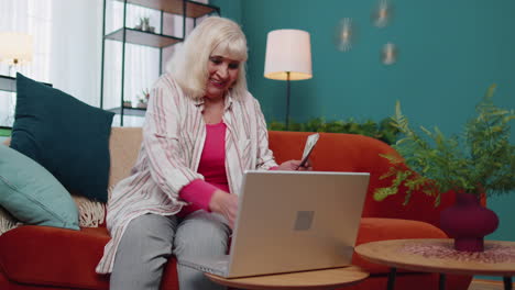 Cheerful-satisfied-elderly-grandmother-freelancer-woman-pressing-button-on-laptop-and-counting-money