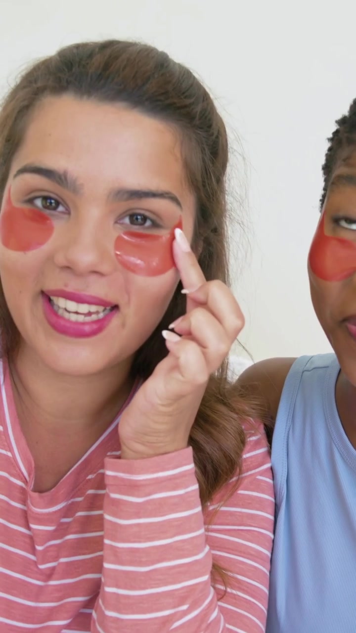 Vertical video: Two women adjusting red-eye patches on bed, pink, blue tops on camera for skincare
