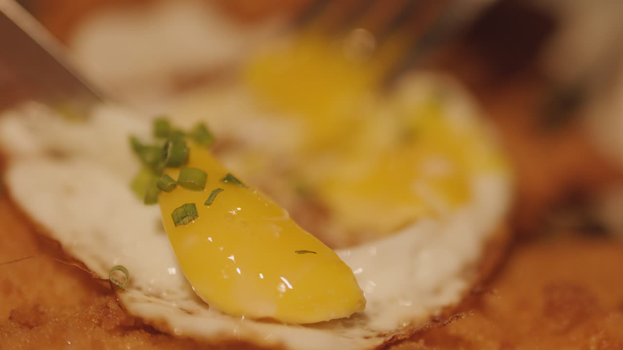 暖かく柔らかな照明の下、黄金色でサクサクのチキンカツの上に、チャイブが添えられた目玉焼きにフォークとナイフが切り込む。