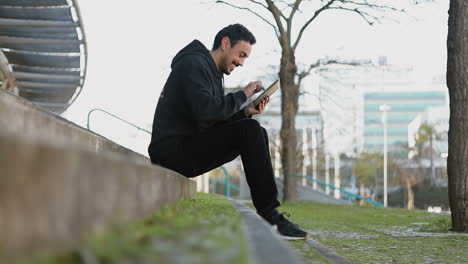 Side-view-of-excited-young-Arabic-handsome-man-with-dark-curly-hair-and-beard-in-black-hoodie-sitting-on-stairs-outside