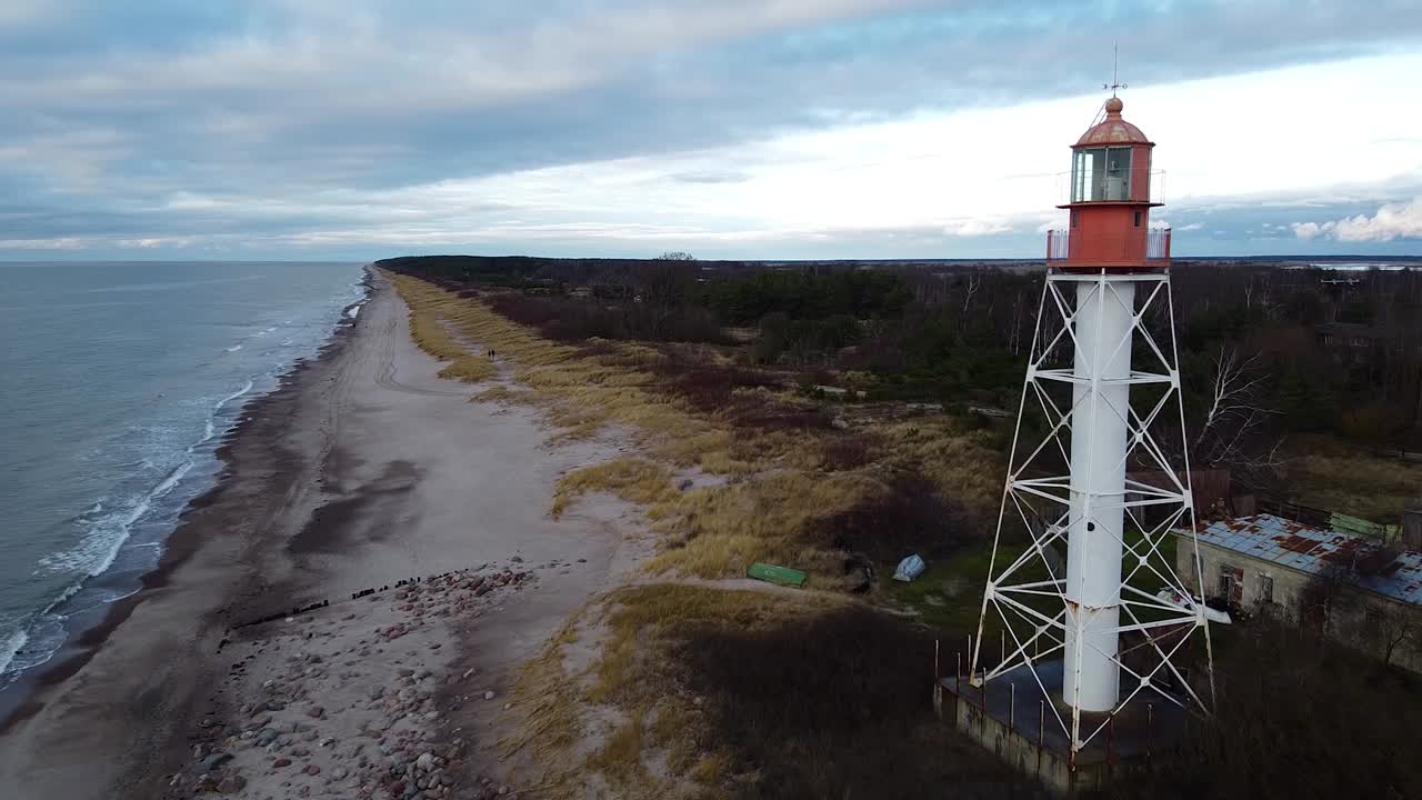 vista aérea del faro de acero pintado de blanco ubicado en pape, letonia en la costa del mar báltico en un día nublado, gran angular que establece disparos de drones moviéndose hacia atrás