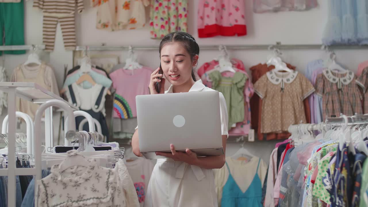 propietaria asiática de una tienda de ropa para niños hablando en su teléfono inteligente mientras usa una computadora portátil para comprobar las existencias en los rieles