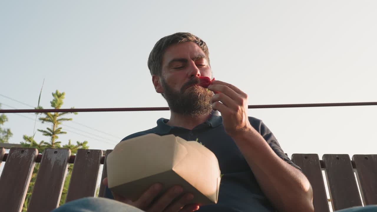 male farmer sitting on swing bench picking fresh strawberry from cardboard container examining bright red fruit under warm backlight then eating fruit with relaxed pose in rural garden