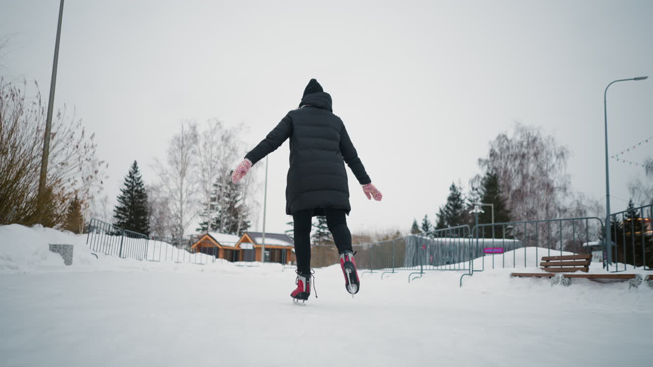 Lady skating on outdoor ice rink in winter surrounded by snow, wearing black coat, knit hat, colorful gloves, red skates, trees and cabins in background, enjoying seasonal activity winter lifestyle