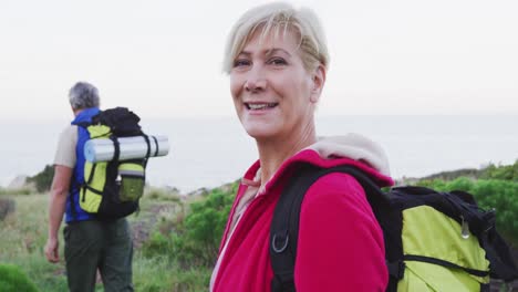 Portrait-of-senior-hiker-woman-with-backpack-and-trekking-poles-smiling-while-standing-on-the-grass-