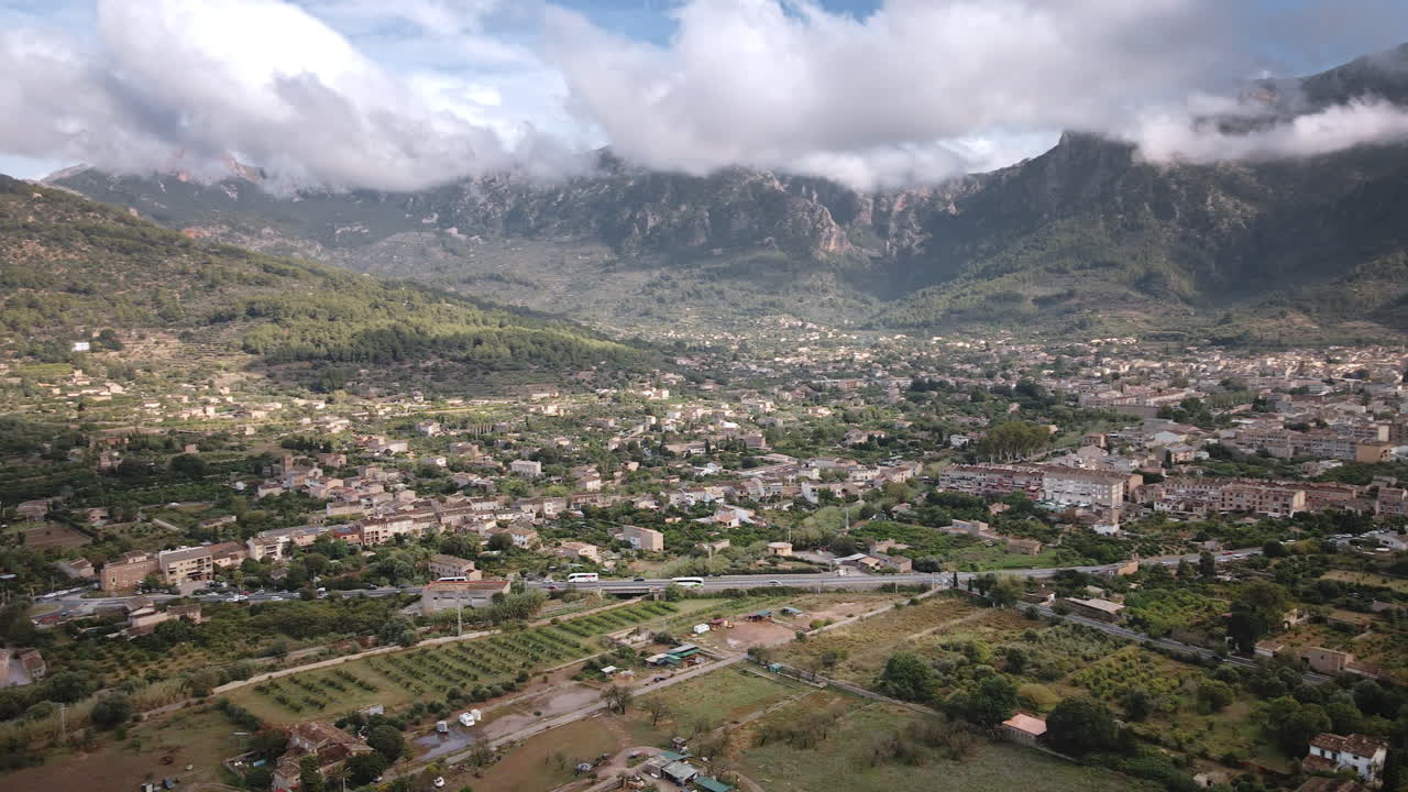 Ripresa aerea da drone con panoramica a sinistra su Sóller, che rivela la cittadina compatta, il fumo dei camini, la valle verde e la Serra de Tramuntana avvolta dalle nuvole in una giornata luminosa