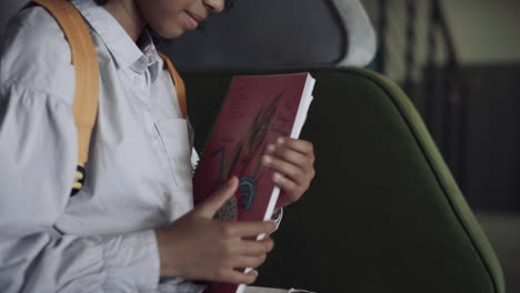 Colegiala-Abriendo-Un-Libro-Sentado-En-Un-Banco-En-El-Pasillo-De-Cerca.-Niña-Africana-Lista-Para-Dibujar.