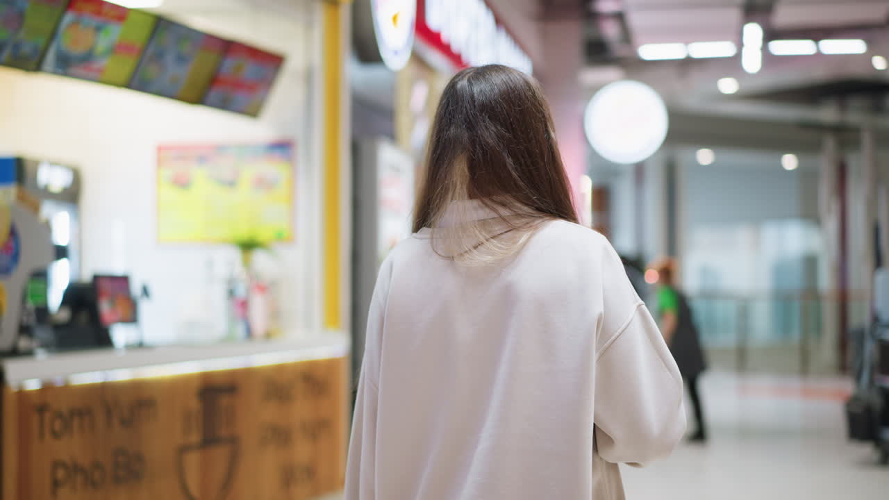 Back view of young lady walking inside mall cafe section wearing casual outfit, blurred view of food stalls and people in background creating modern lifestyle atmosphere with bright indoor lighting