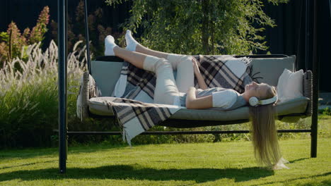 a teenage girl listens to music on headphones, lies on a garden swing in the backyard of the house.