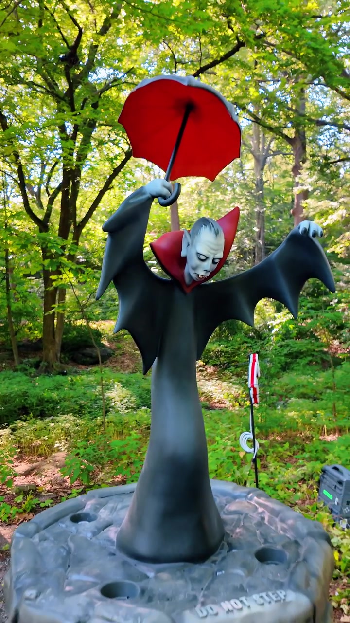 New York, USA, 17 August 2025: Character sculpture of vampire with red umbrella in green forest. Vampire figure with wings and red umbrella surrounded by lush green trees in a park.