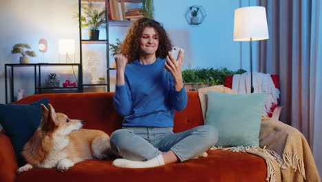 surprised woman browsing smartphone celebrating success winning lottery sitting beside corgi dog