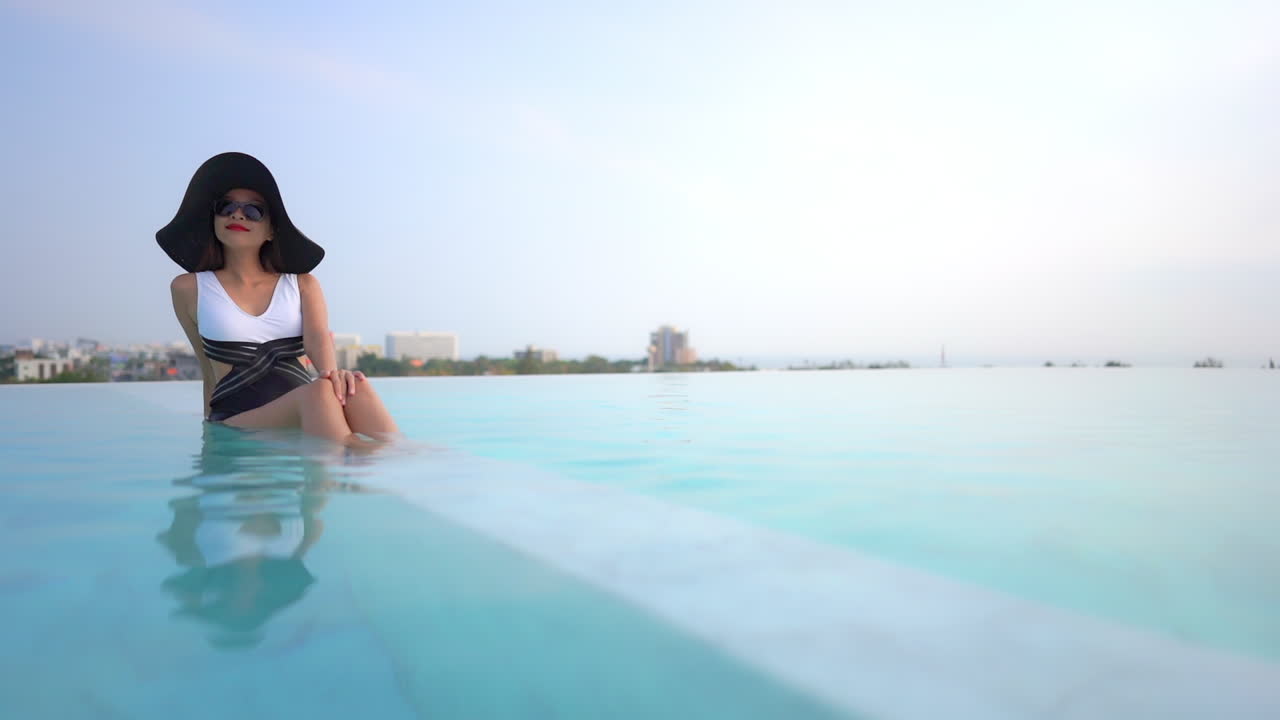 dama en traje de baño y sombrero grande relajándose en una piscina cálida y clara con vistas a los edificios, el paisaje y el mar en el fondo
