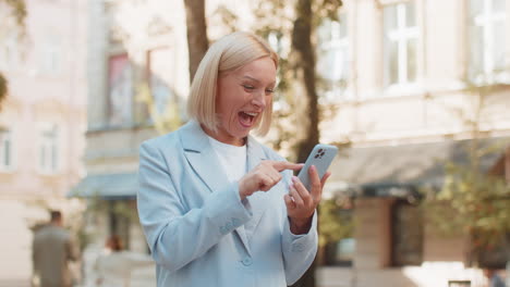 mature caucasian businesswoman ceo celebrating good news on smartphone standing on city street